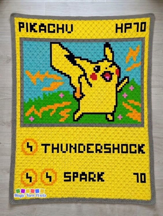 A finished Pikachu Pok&eacute;mon Card C2C crochet blanket is displayed flat on a light wood floor. The blanket features Pikachu inside a Pok&eacute;mon trading card layout with "PIKACHU," "HP70," lightning effects, and the attacks "THUNDERSHOCK" and "SPARK," bordered in gray. Original design by Ariana of MagicYarnPixels.