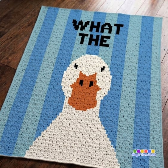 Finished C2C crochet blanket laid on a wooden floor, showing a large white duck with an orange bill against blue striped columns. Bold black text reading "WHAT THE" appears above the duck. Original design by Ariana of MagicYarnPixels.