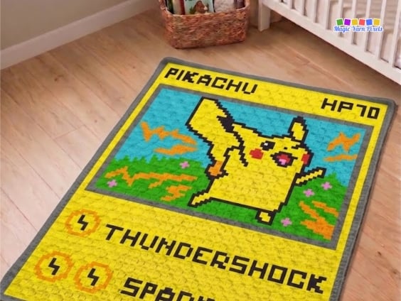 Crochet Pikachu trading card blanket displayed on a light wooden floor in a nursery beside a white crib. The corner-to-corner (C2C) blanket has a gray border and features a pixel-style Pikachu inside a trading card design with a blue sky, green grass, orange lightning bolts, and the words "PIKACHU," "HP70," "THUNDERSHOCK," and "SPARK." Original design by Ariana of MagicYarnPixels.