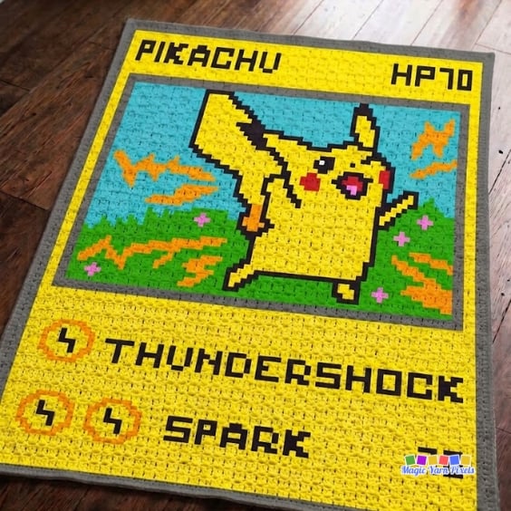 Corner-to-corner (C2C) crochet Pikachu trading card blanket spread across a dark wooden floor. The blanket shows a pixel-style Pikachu framed inside a yellow trading card with a gray border, blue sky, grassy field, orange lightning bolts, and the text "PIKACHU," "HP70," "THUNDERSHOCK," and "SPARK." Original design by Ariana of MagicYarnPixels.